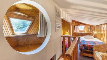 A cozy cabin room with a circular skylight, a bed with a plaid blanket, a wooden chair, and a small table with a lamp.