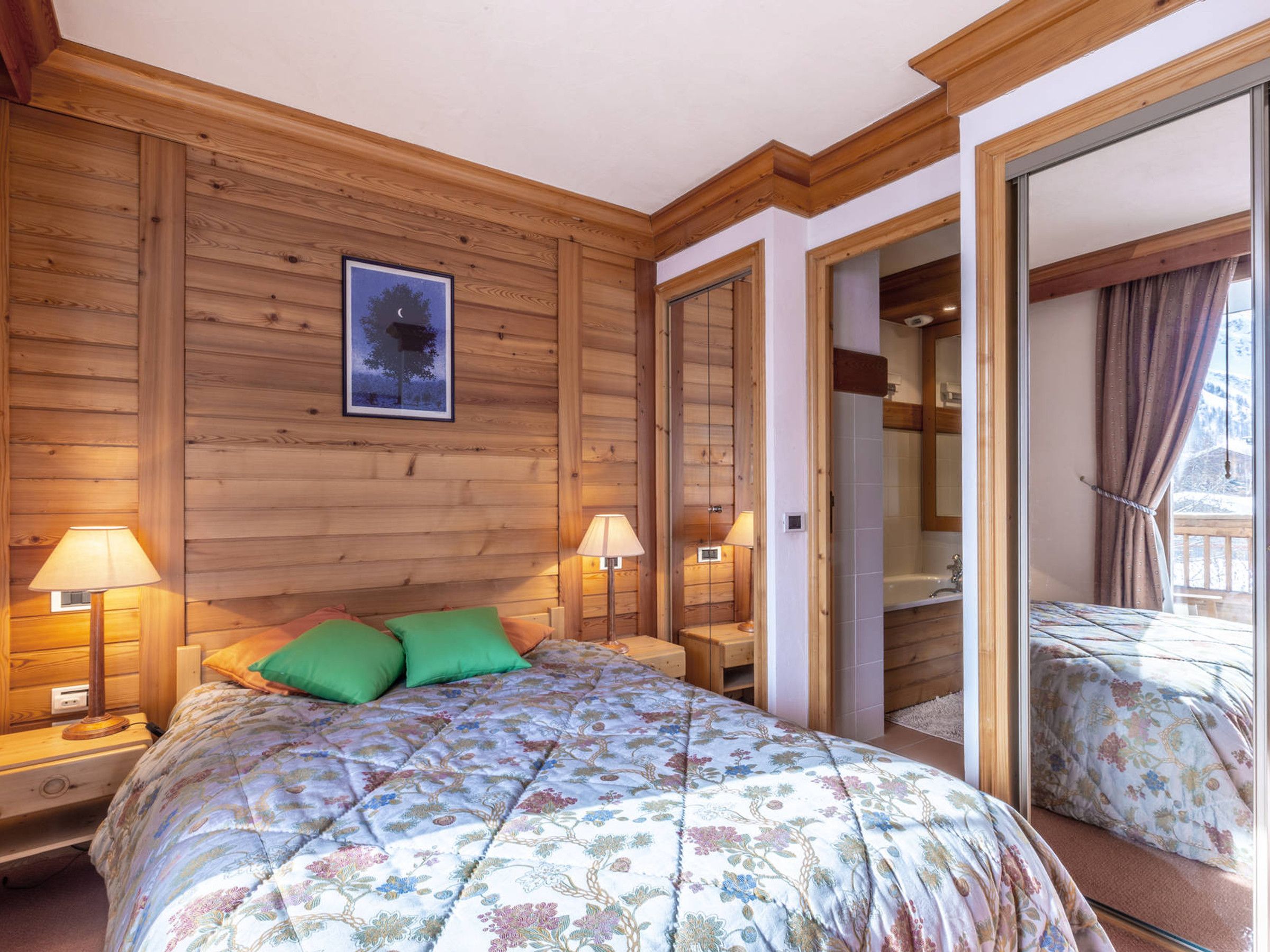 Ein Zimmer mit Holzboden, einem Bett, zwei Nachttischlampen und einem gerahmten Bild an der Wand.
