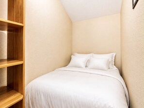 A small, neatly arranged bedroom with a single bed, a wooden shelf unit, and a light-colored wall.