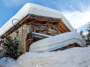 A snow-covered chalet with a stone facade, wooden beams, and a wooden staircase leading to a balcony.
