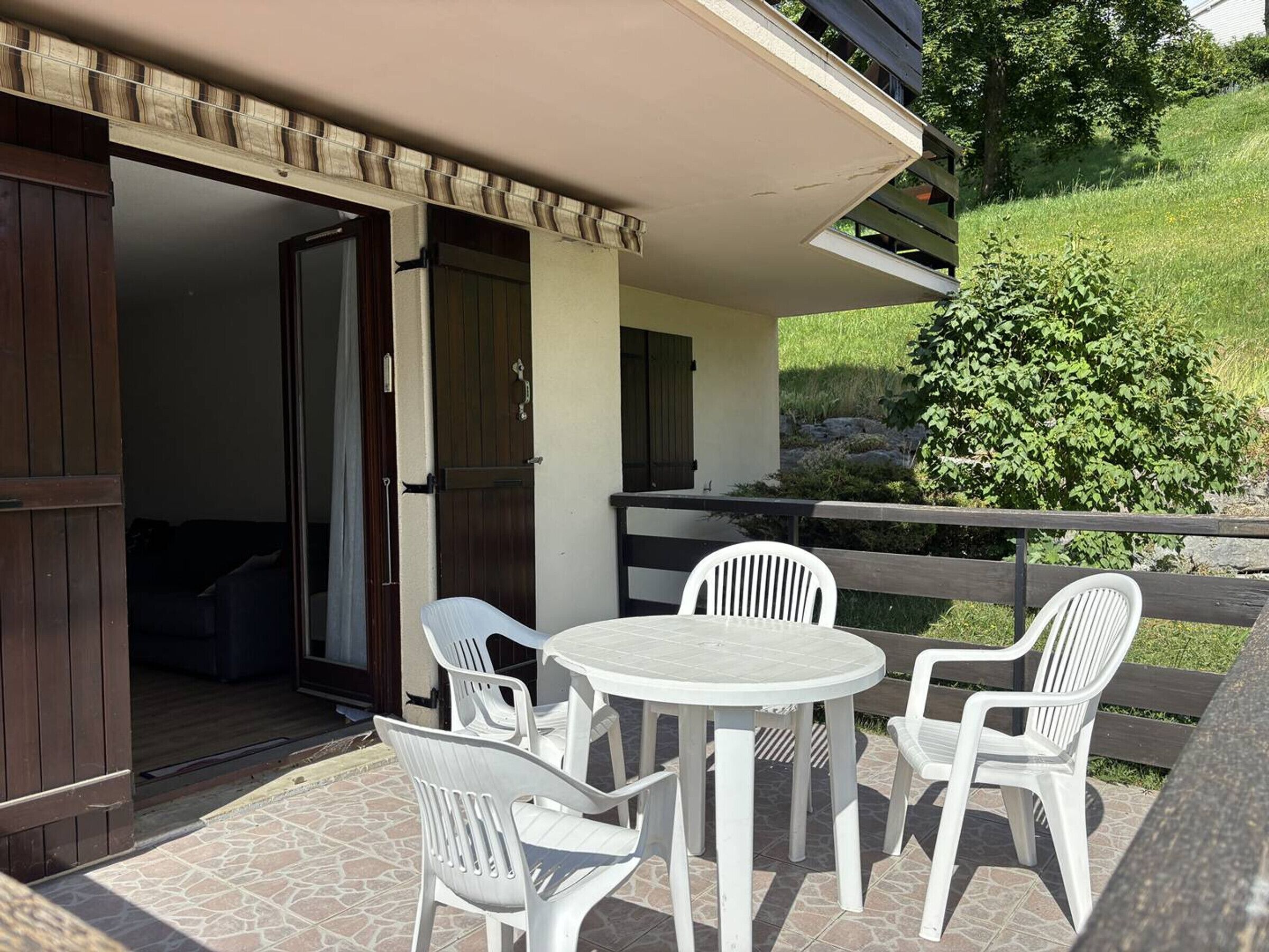 Une terrasse avec une table ronde et quatre chaises blanches, une clôture en bois et un bâtiment dont les portes sont ouvertes.
