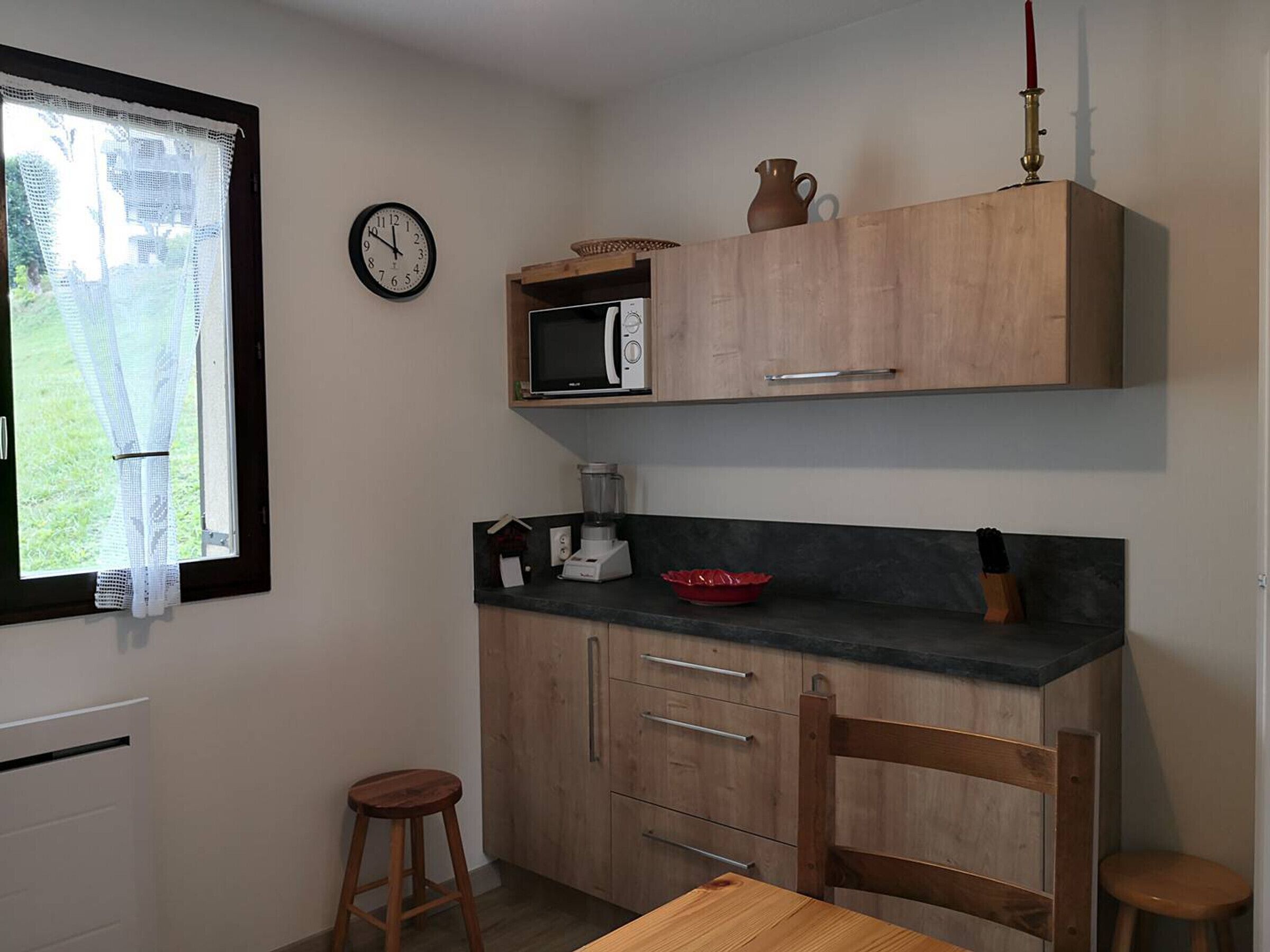 Une petite cuisine équipée d’un four à micro-ondes, d’une machine à café et d’un saladier rouge posé sur le plan de travail. On trouve un meuble en bois au-dessus du plan de travail et une horloge au mur.