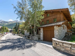 A stone building with a wooden facade, a garage, and a driveway leading to a street lined with trees and other buildings. - Luxury Duplex Apartment in Central Val-d'Isère, Near Slopes, with Large South-Facing Terrace (Val-d'Isère)