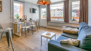 Una sala de estar moderna con sofá, mesa de comedor y sillas. Hay un televisor montado en la pared y una ventana con cortinas.