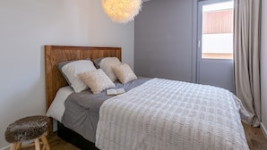 Un dormitorio con cabecera de madera, ropa de cama gris, una lámpara blanca y mullida, y un taburete pequeño.