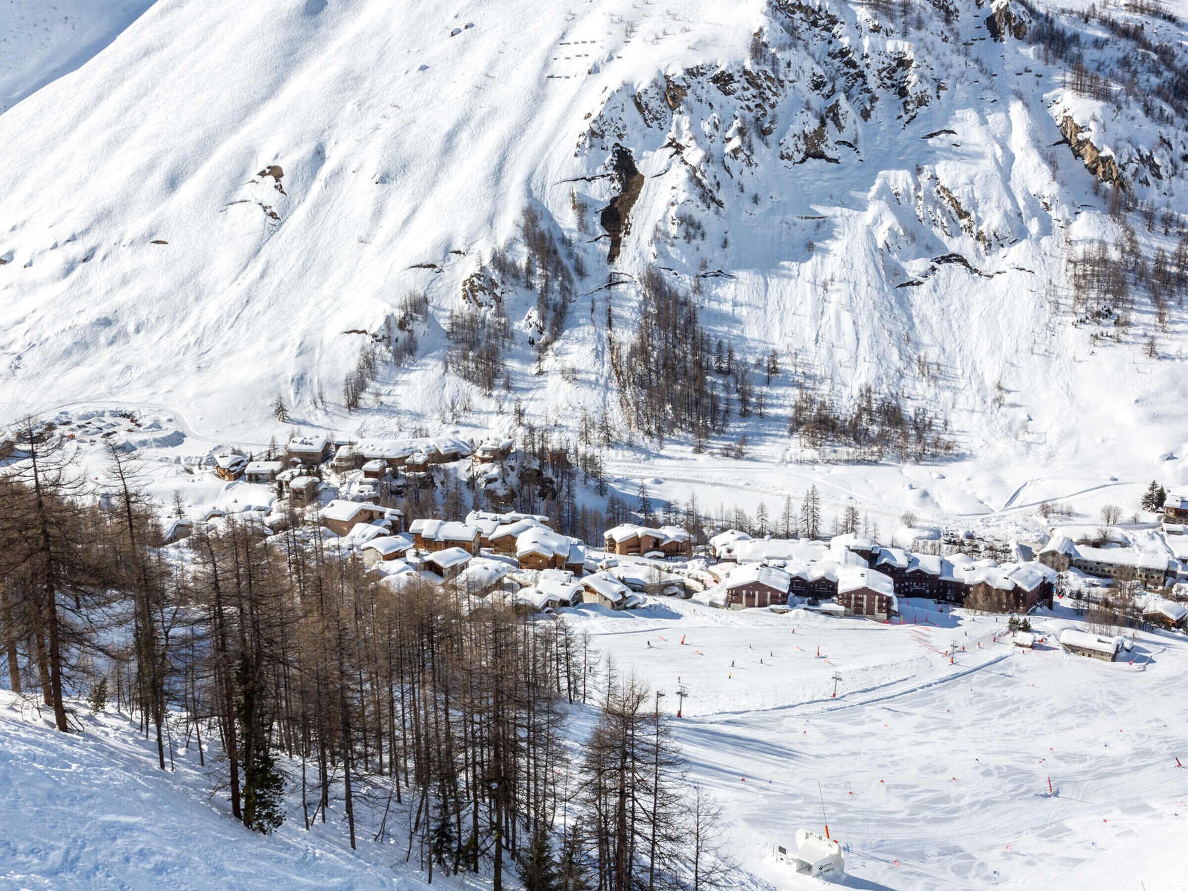 A snow-covered mountain village with traditional wooden houses clustered together, surrounded by trees and a ski slope.
