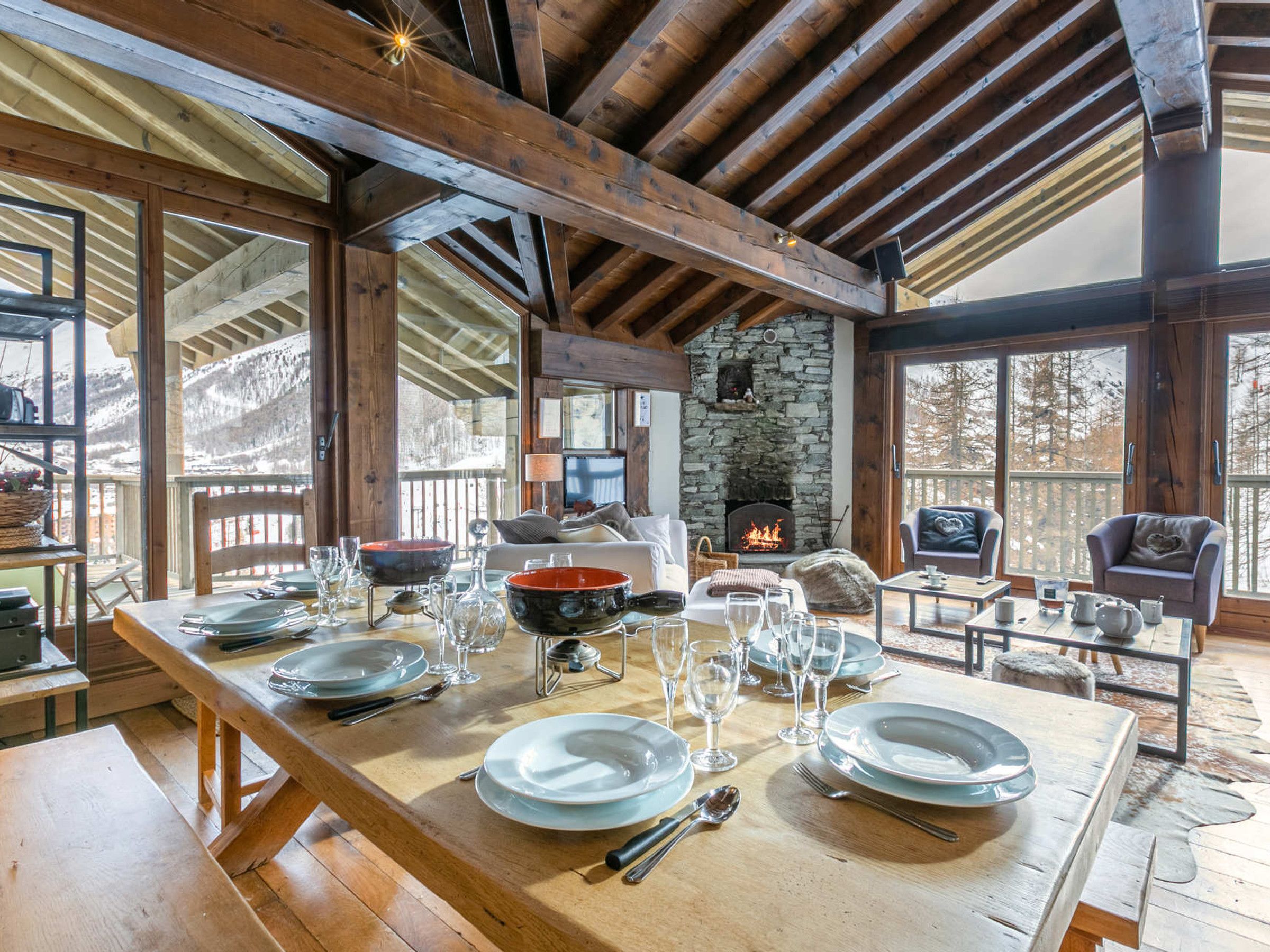 A wooden dining table set for four with plates, glasses, and cutlery. A stone fireplace is visible in the background.