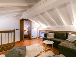 A loft-style bedroom with a sloped ceiling, wooden beams, a bed with red pillows, a small round table, and a green sofa with a fur throw.