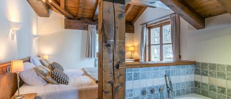 Une chambre avec un plafond en bois, une grande fenêtre, un lit avec du linge de maison blanc et une salle de bain avec un mur carrelé et une baignoire.