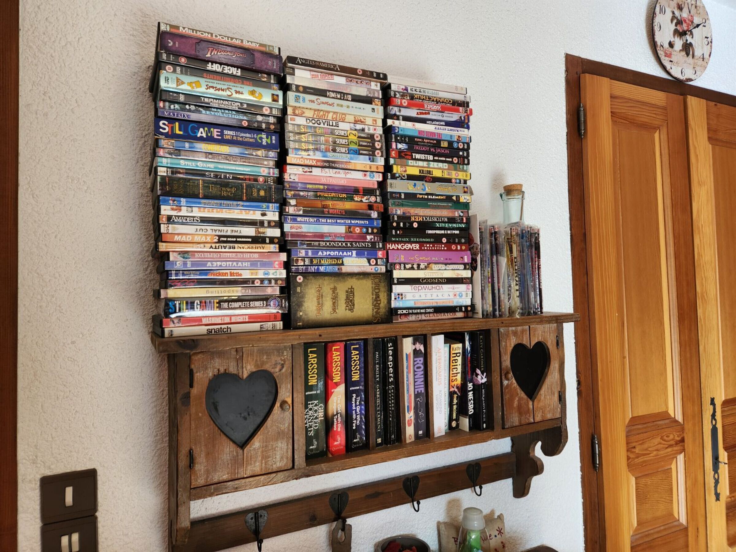 A collection of DVDs and books on a wooden shelf against a wall, with a clock above and a wooden door to the right.