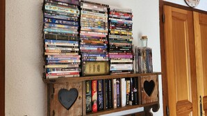 A collection of DVDs and books on a wooden shelf against a wall, with a clock above and a wooden door to the right. - T2 apartment with mezzanine in village center, pets allowed (Samoëns)