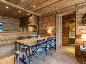 A rustic wooden interior with a dining table and chairs, a kitchen area with a sink and stove, and a bathroom visible through an open doorway.