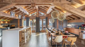 A spacious kitchen with a high ceiling and exposed wooden beams, a central island with stools, and a dining area with a long table and chairs.