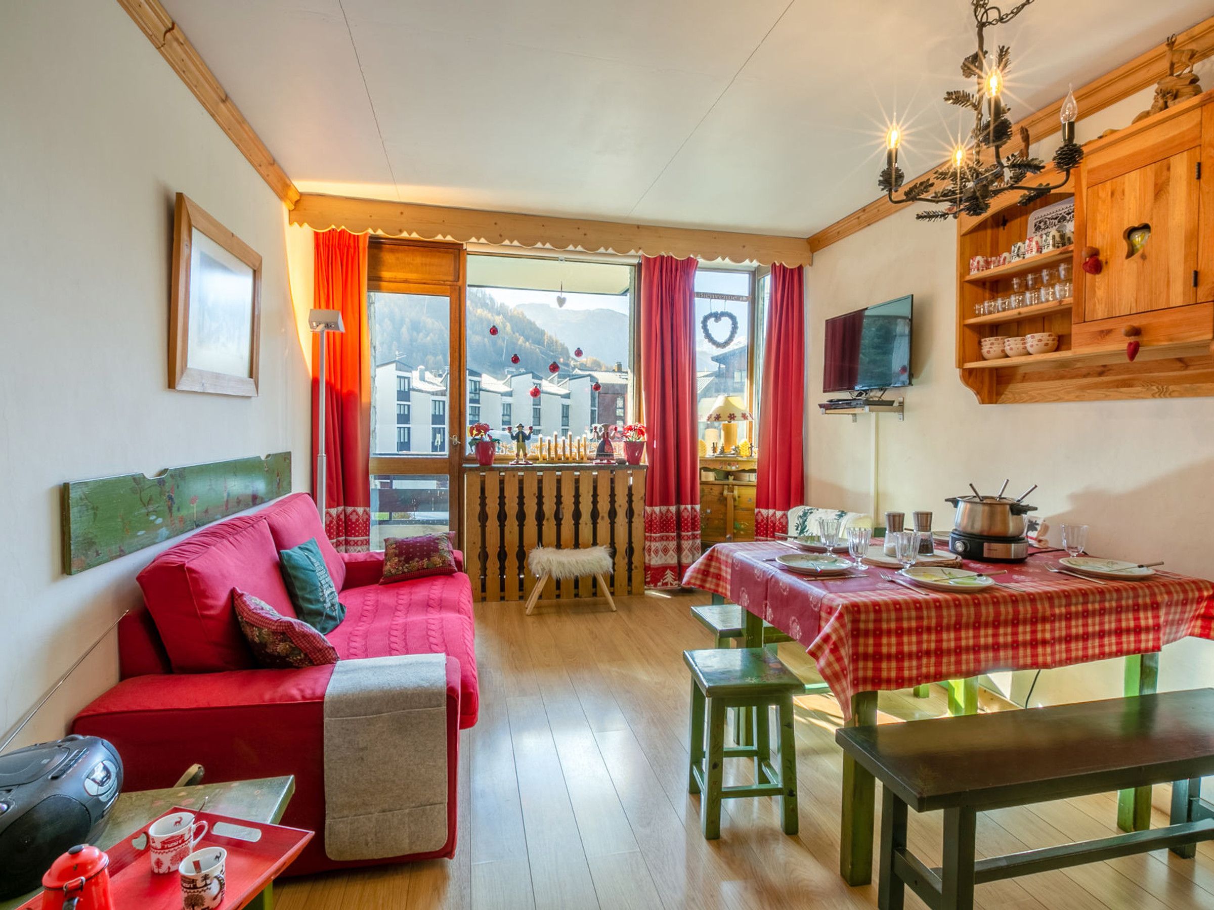 A cozy living room with a red sofa, wooden dining table, and a view of a mountain village through large windows.
