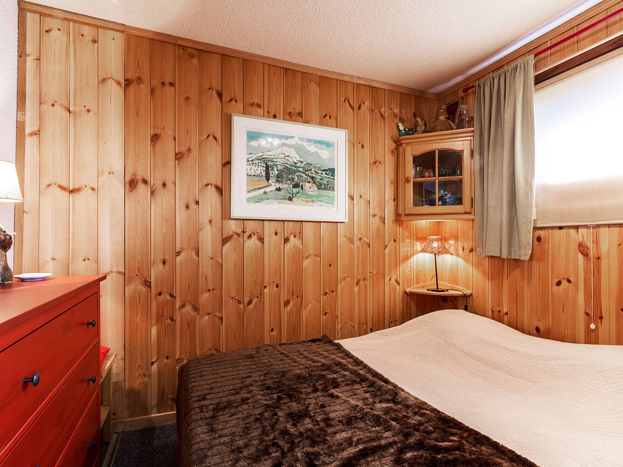 A room with wooden panel walls, a bed with a dark brown blanket, a red dresser, a framed picture on the wall, a cabinet with glass doors, and a lamp on a bedside table.
