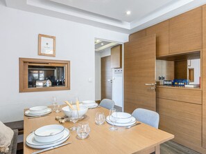 A dining area with a wooden table set for four, a kitchen visible through a window, and a wooden cabinet with a mirror.