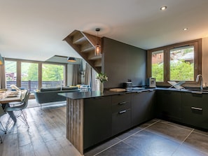 A modern kitchen with a central island, dark cabinetry, and large windows offering a view of greenery.