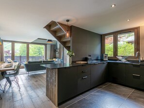 A modern kitchen with a central island, dark cabinetry, and large windows offering a view of greenery.