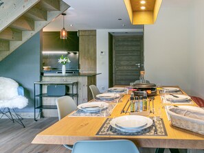 Un espace repas moderne avec une table en bois dressée pour quatre personnes, une cuisine en arrière-plan et un escalier menant à un autre niveau.