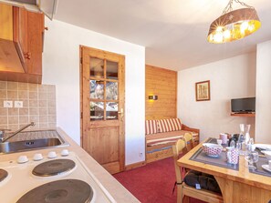 A compact kitchen with a stove, sink, and wooden cabinets. There's a wooden door with a window, a bed with striped pillows, and a TV on the wall.