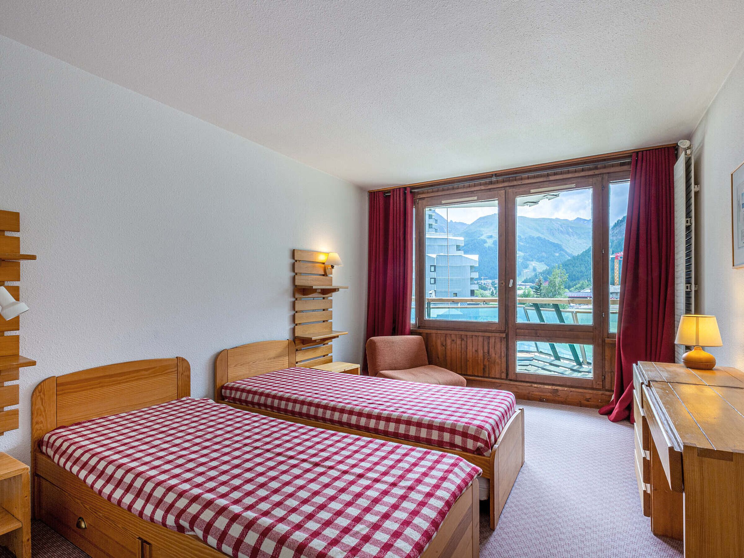 Une chambre avec deux lits en bois, recouverts d’une literie à carreaux, une table de chevet en bois, une lampe, une fenêtre donnant sur la montagne et un rideau rouge.