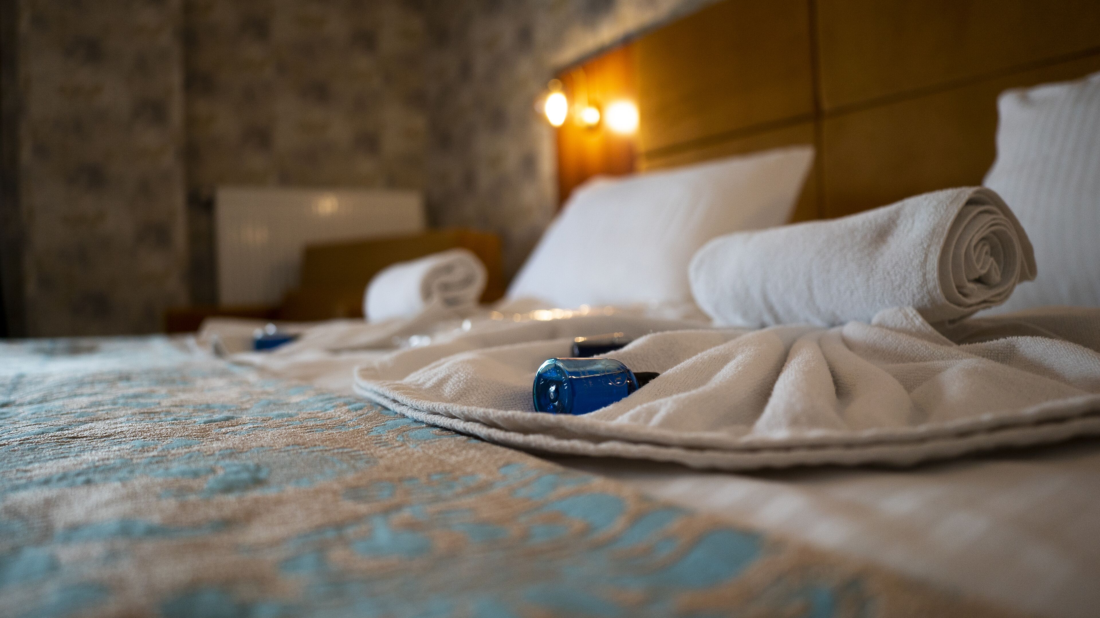 A neatly made bed with rolled towels and a blue bottle, suggesting a hotel room setting.