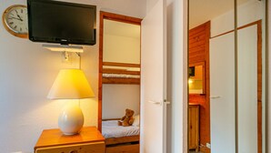 Una habitación con tocador de madera, lámpara de noche, cama con un osito de peluche, televisor montado en la pared y un espejo.