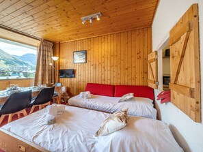 A hotel room with wooden panel walls, a bed with red bedding, a TV mounted on the wall, and a dining area with a table and chairs.