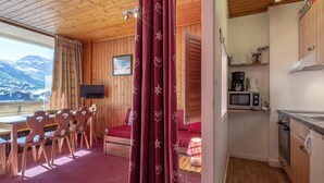 Une pièce chaleureuse avec un plafond en bois, un coin repas avec des chaises et une table, un lit recouvert d’une couverture rouge, une cuisine équipée d’un four à micro-ondes et d’une machine à café, et une fenêtre donnant sur les montagnes.
