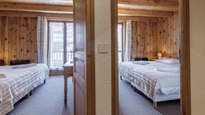 A wooden cabin room with two beds, a desk, and a window with curtains.