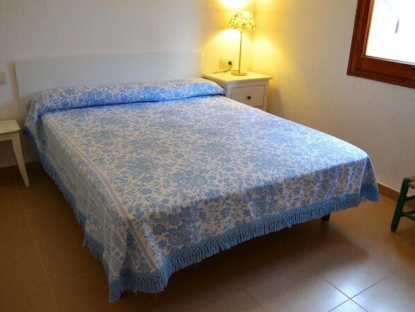 A bed with a blue floral bedspread, a bedside table with a lamp, and a window with a wooden frame. - Family ground floor in Cap de Ras, pets allowed (Llançà)