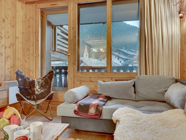 A cozy cabin interior with a sofa, a chair, a table with mugs, and a view of a snowy village through large windows.