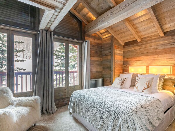 A wooden cabin bedroom with a bed, a chair, and a view of a snowy landscape through large windows.