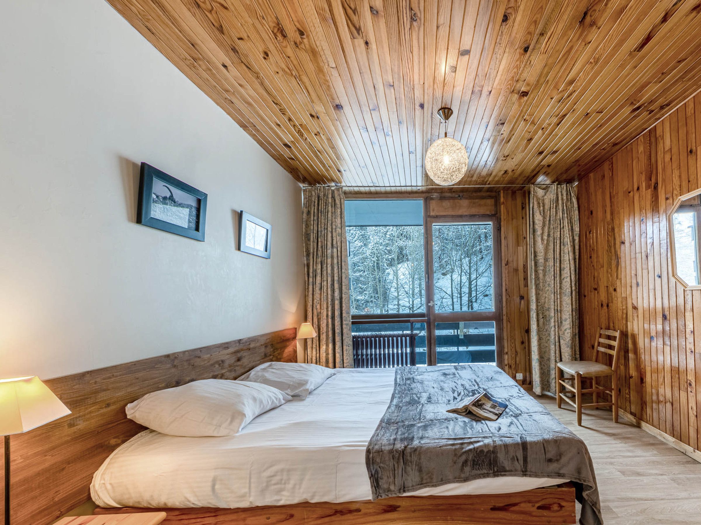 A wooden-paneled bedroom with a bed, a reading lamp, a chair, and a window with curtains.