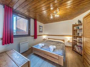 A bedroom with a wooden ceiling, a bed with a headboard, a bedside table, a bookshelf, and a window with curtains. - Charming Apartment with South-Facing Balcony in Heart of Resort, Near Ski Slopes (Val-d'Isère)