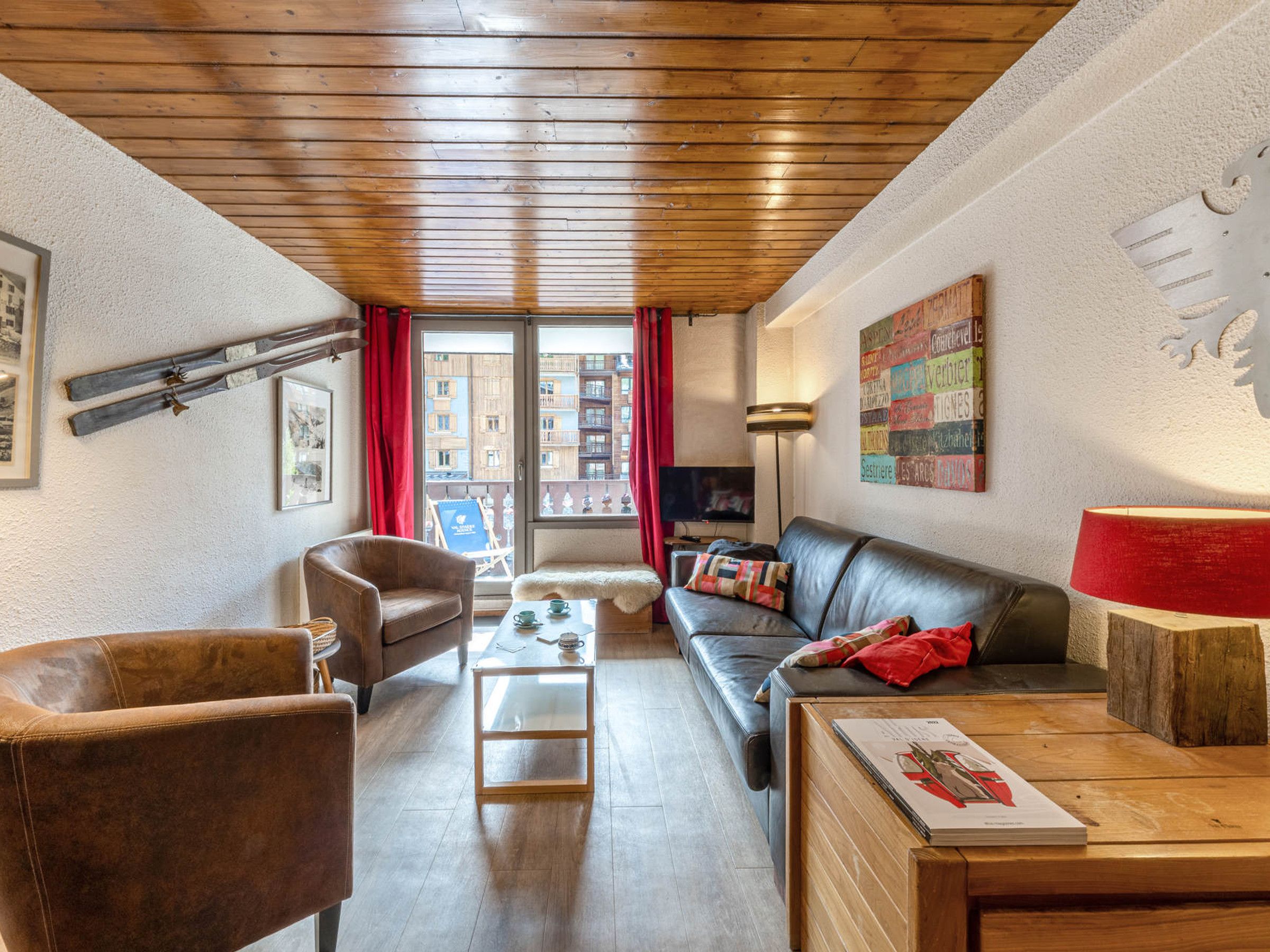 Een gemeubileerde woonkamer met een bank, fauteuil en salontafel, een houten plafond en stadszicht door een raam.