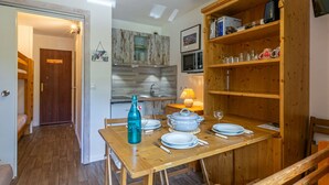 A dining area with a wooden table set for four, a kitchen in the background, and a wooden bookshelf with various items.