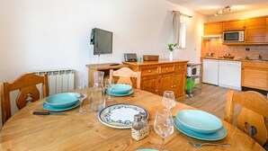 Ein Essbereich mit einem Holztisch, gedeckt für vier Personen, umgeben von Stühlen. Im Hintergrund ist eine Küche mit Holzschränken und Geräten zu sehen.