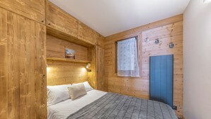 A bedroom with a wooden headboard, a bed with white linens and a gray blanket, a small window with a sheer curtain, and a blue door.