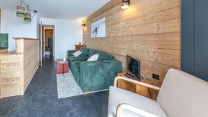 A modern living room with a green sofa, a wooden coffee table, and a flat-screen TV. There is a wooden wall paneling and a fireplace.