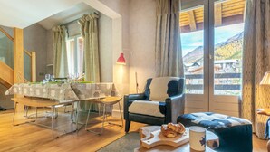 Ein gemütliches Wohnzimmer mit Esstisch für zwei Personen, einem bequemen Sessel, einem Holztisch mit Gebäck und Blick auf eine Berglandschaft durch große Fenster.