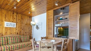 A cozy room with wooden walls, a wooden sofa with a checkered cushion, a dining table with chairs, and a kitchen area with wooden cabinets and a tiled backsplash.