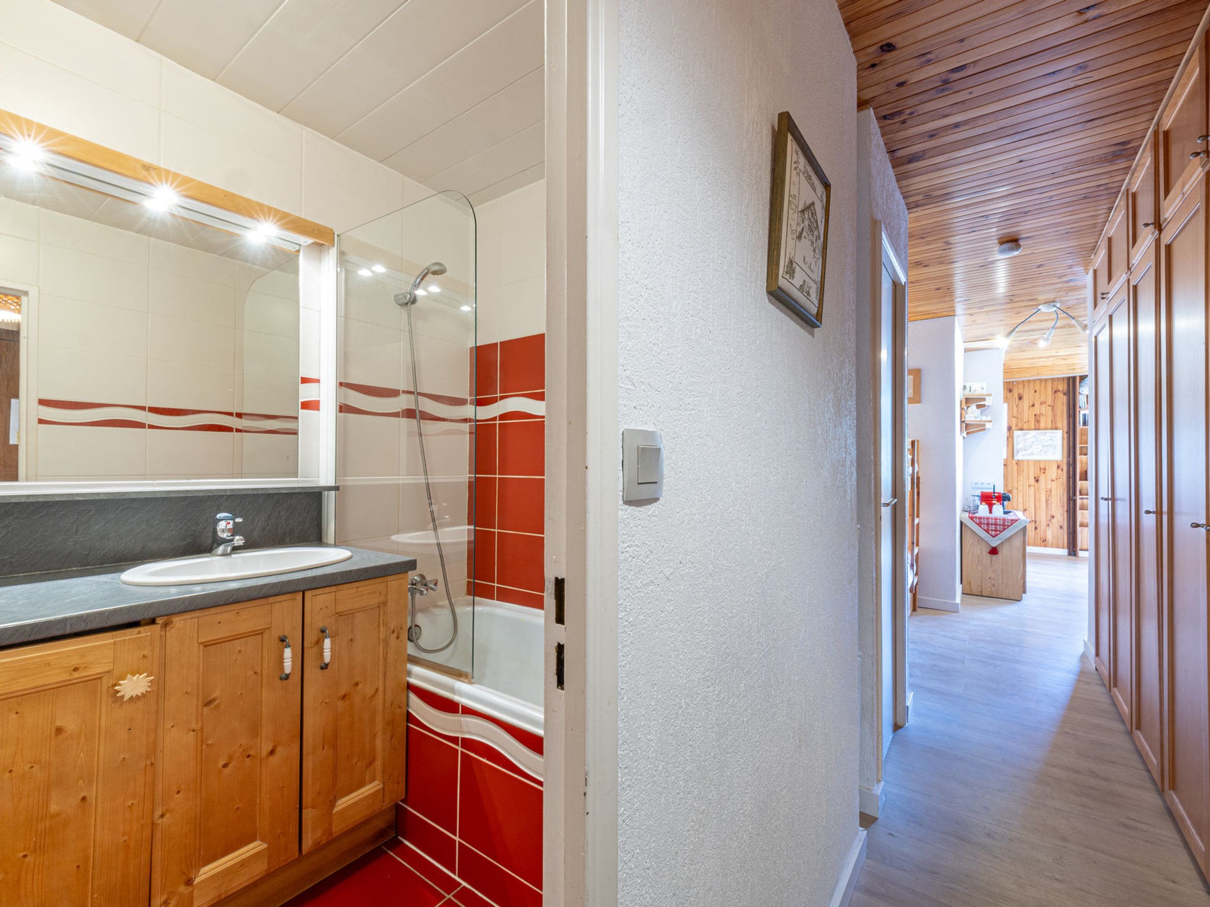 Un baño con tocador de madera, cabina de ducha con mampara de vidrio y pasillo que conduce a una cocina.