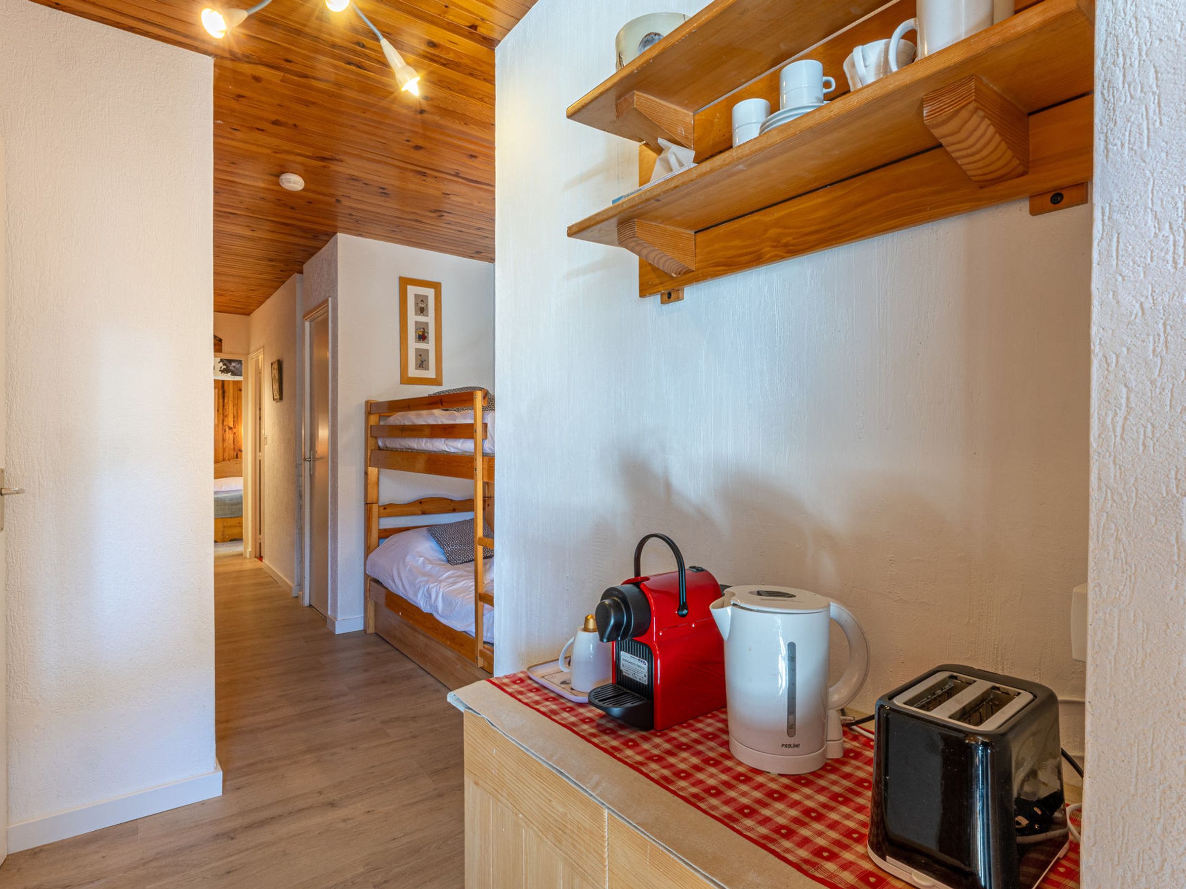 Un couloir étroit avec un plancher en bois, un lit superposé, une petite cuisine équipée d’une bouilloire rouge, d’une machine à café blanche et d’un grille-pain.