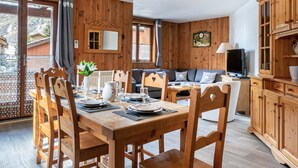 A wooden-furnished dining area with a table set for four, a sofa, and a television in a room with wooden walls and a large window. - Rustic Mountain-Style Apartment in Chalet, Village Center, Ski-In/Ski-Out (Val-d'Isère)
