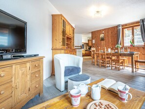 Een gezellige woonkamer met een flatscreen televisie, houten kasten, een witte fauteuil en een eethoek met houten meubels.