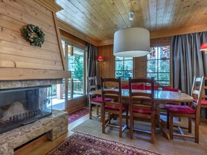 A wooden interior with a fireplace, a dining table with chairs, and large windows.