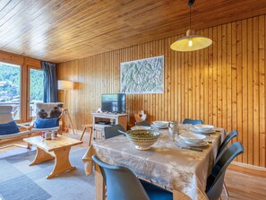 Ein Zimmer mit Holzwandverkleidung, einem Esstisch für vier Personen, einem Fernseher und einem mit Vorhängen drapierten Fenster.