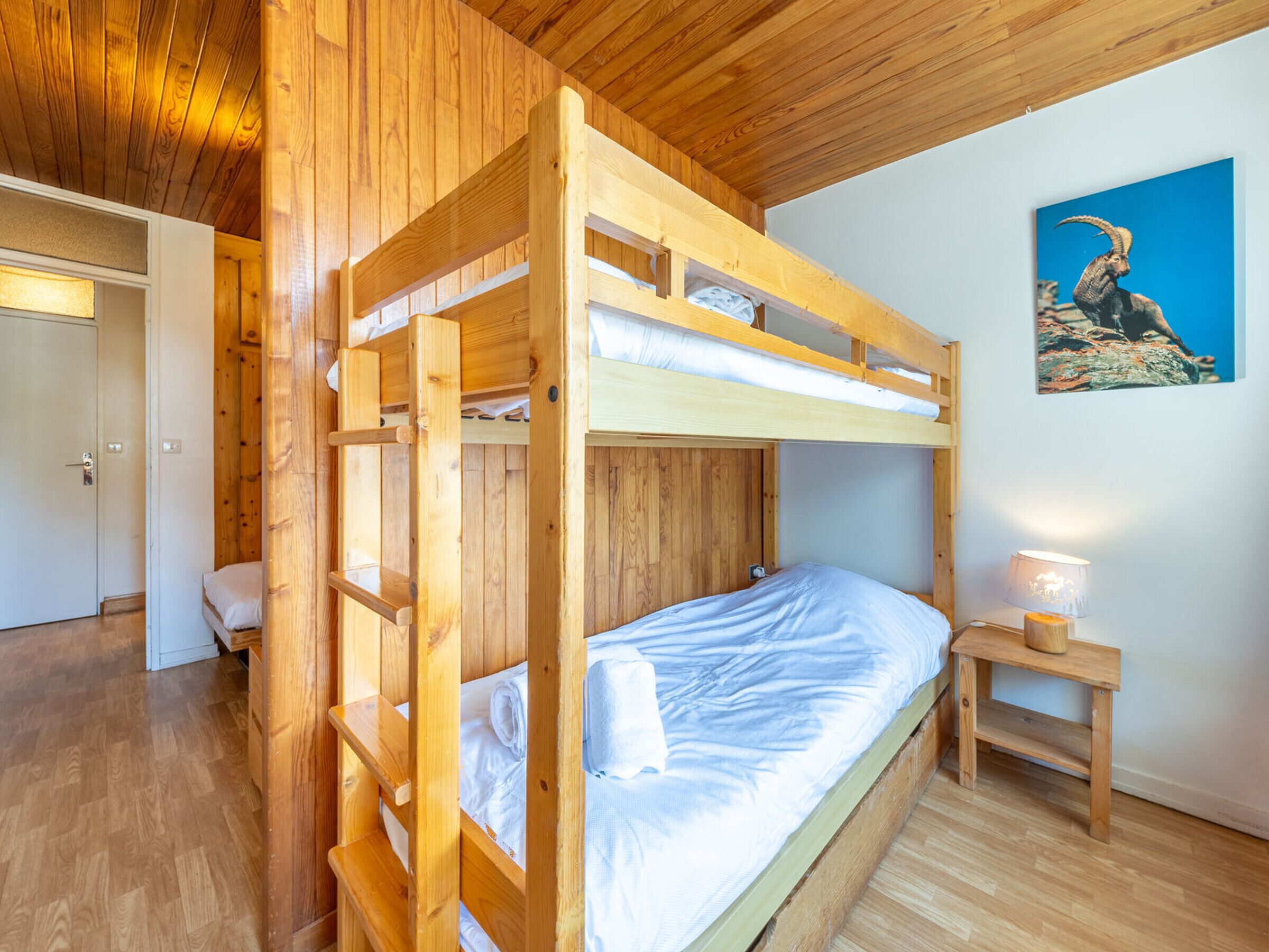 A bunk bed room with wooden walls and a ceiling, a small bedside table with a lamp, and a painting on the wall.