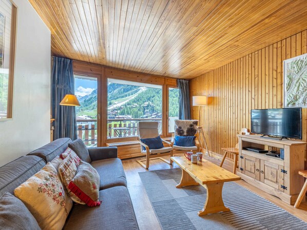 Una sala de estar acogedora con sofá, sillones, una mesa de centro y televisión. Amplias ventanas ofrecen vistas a la montaña.